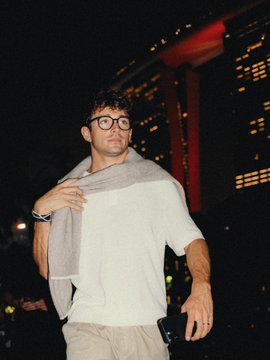 A man wearing round glasses, a white long-sleeved shirt, and a gray sweater draped over his shoulders stands outdoors at night in front of a red-lit building. The accompanying caption jokes that his name initially made him look like a Binance KOL.