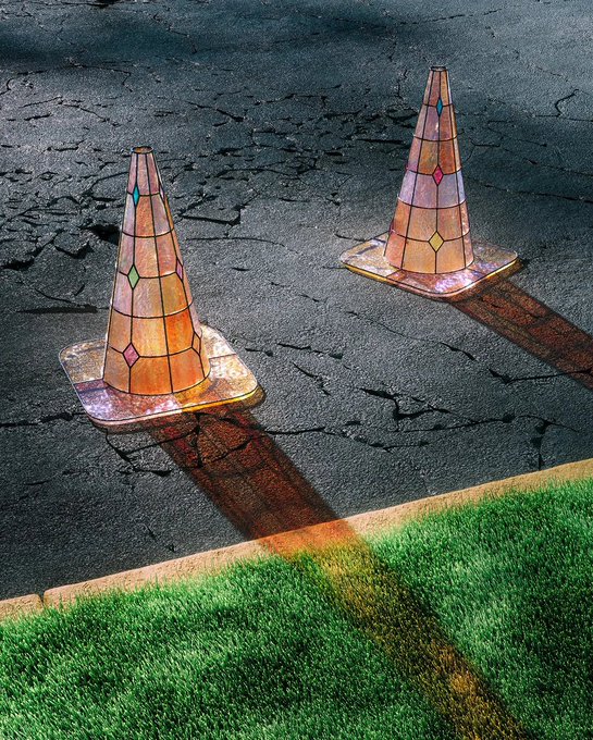 Two translucent traffic cones with a stained-glass pattern cast colorful shadows on cracked asphalt next to a strip of artificial grass.