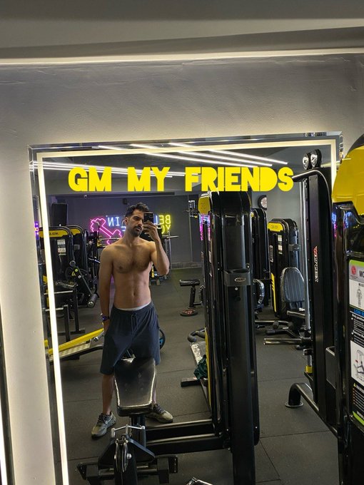 A shirtless man takes a gym selfie with the text 'GM MY FRIENDS' overlaid in yellow.