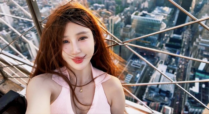 A young woman with long auburn hair taking a selfie on a high observation deck with a cityscape visible in the background.
