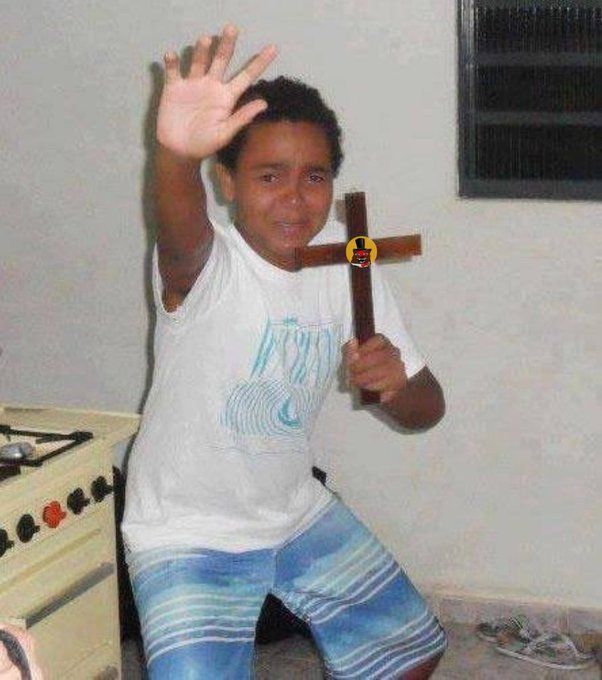 A young boy in a white t-shirt and blue shorts sitting indoors, holding up one hand and gripping a wooden cross with the other.