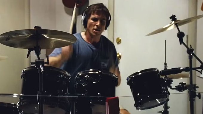 A young man wearing headphones plays a black drum set in what appears to be a practice room.