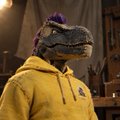 A person wearing a dinosaur mask and a yellow hoodie stands in a warmly lit indoor setting.