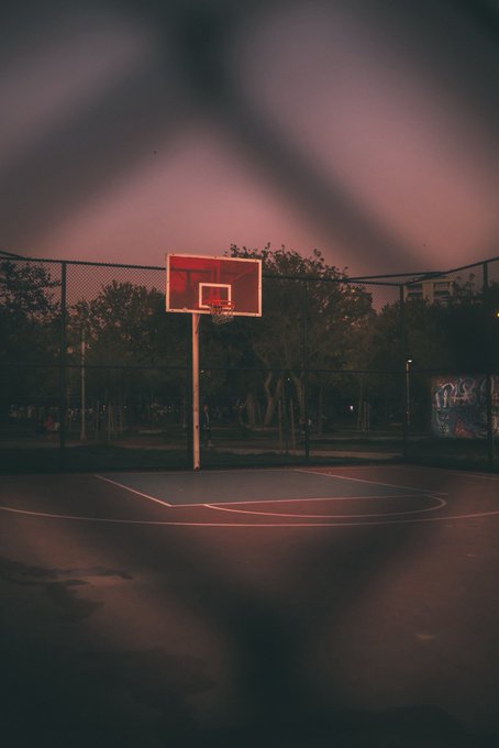 A moody, low-light photograph of an empty outdoor basketball court at dusk, framed through a chain-link fence with a hoop visible in the center.