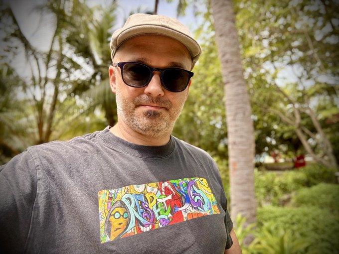 A man wearing a beige cap, dark sunglasses, and a gray t-shirt with colorful artwork stands outdoors in a tropical setting with palm trees.