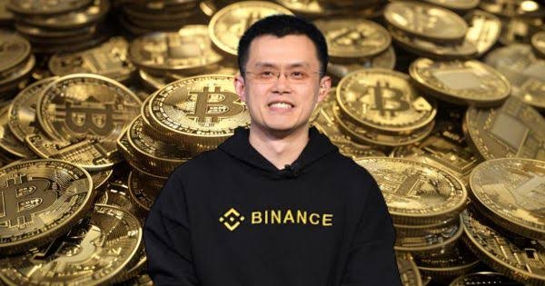 Binance founder Changpeng Zhao smiling while wearing a black Binance hoodie against a backdrop of physical Bitcoin coins.