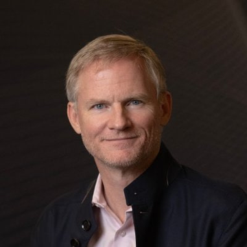 A portrait of a middle-aged man with short blonde hair, wearing a dark blazer over a white shirt, pictured here as BitGo CEO Mike Belshe in connection with his stance rejecting AI regulation proposals.