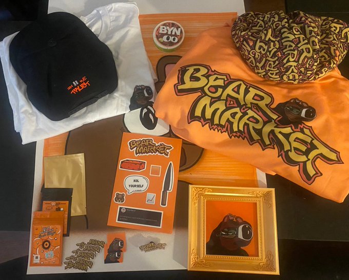 A flat lay of Bear Market merchandise including a 'WE TRUST' cap, an orange t-shirt, a sticker sheet, a pin, and an NFT card with a bear character.