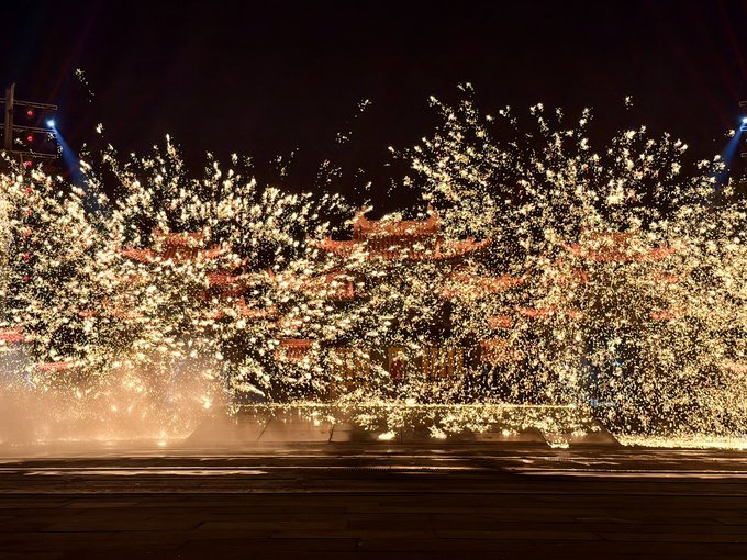 A spectacular nighttime display of molten sparks or fireworks bursting outward in a massive spray of golden light against a dark sky.