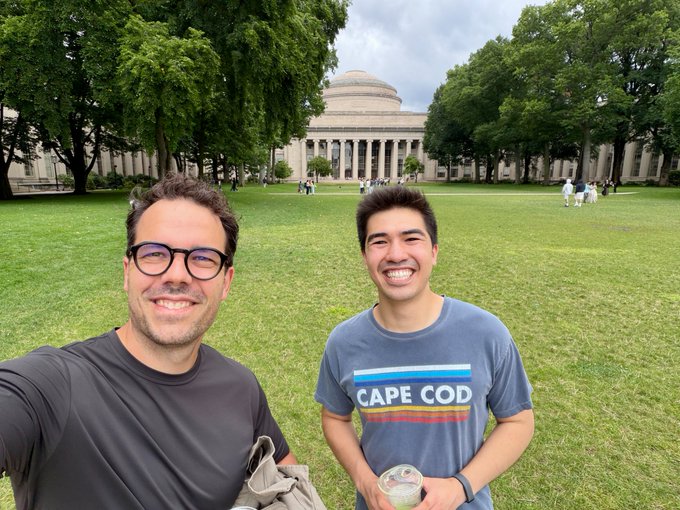 A man takes a selfie with another smiling young man wearing a "CAPE COD" t-shirt while holding a coffee drink, posing on the grassy grounds in front of MIT's iconic domed Great Dome building. The image visually documents the pair's coffee walk to MIT as referenced in the caption.