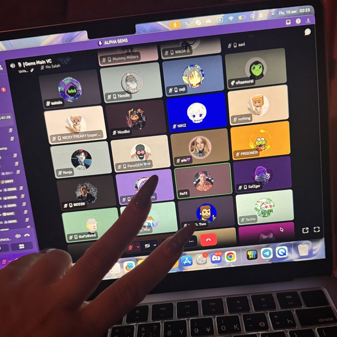 A laptop screen showing a Discord server called 'ALPHA GEMS' with a grid of user avatars and names, as someone's fingers reach up in a peace sign gesture toward the display.