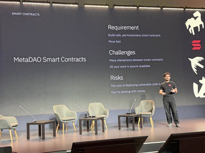 A presenter stands on stage during a presentation about "MetaDAO Smart Contracts," with a slide listing requirements, challenges, and risks of building permissionless smart contracts.