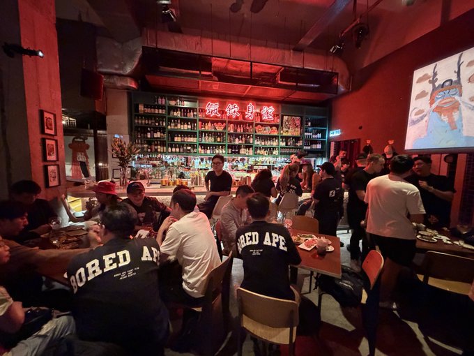 A crowded, dimly lit bar with neon signs displaying Chinese characters ("銀梁身館"), where patrons wearing black "BORED APE" shirts gather around tables, connecting to the post's celebration of meeting the BAYC (Bored Ape Yacht Club) and MAYC (Mutant Ape Yacht Club) community.