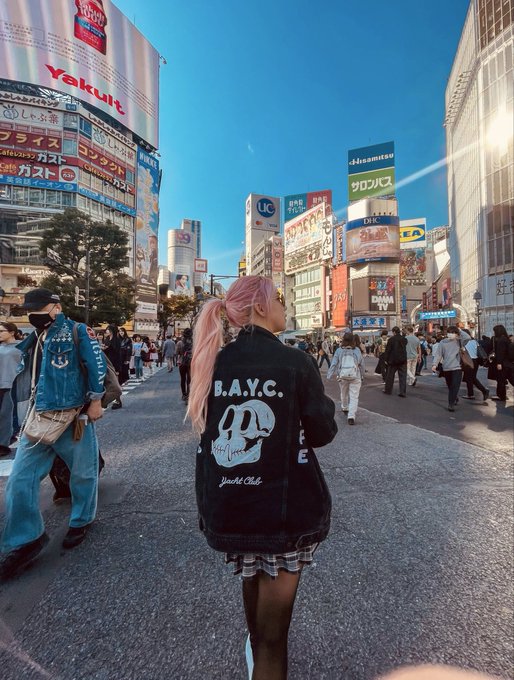 A person with pink hair wearing a black "B.A.Y.C." hoodie with a cartoon face design walks through a busy Japanese shopping district, with "Yakult" and other signs visible in the background.