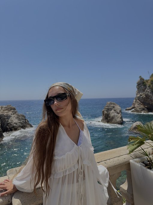 A woman with long hair wearing a white flowy dress, sunglasses, and a headscarf, posing by a stone railing with a rocky coastline and blue sea in the background.