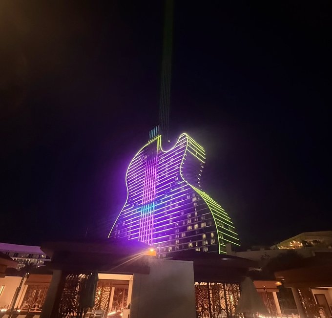 The Hard Rock Hotel in Hollywood, Florida at night, its guitar-shaped building illuminated in purple and green neon lights with a beam shining into the sky.