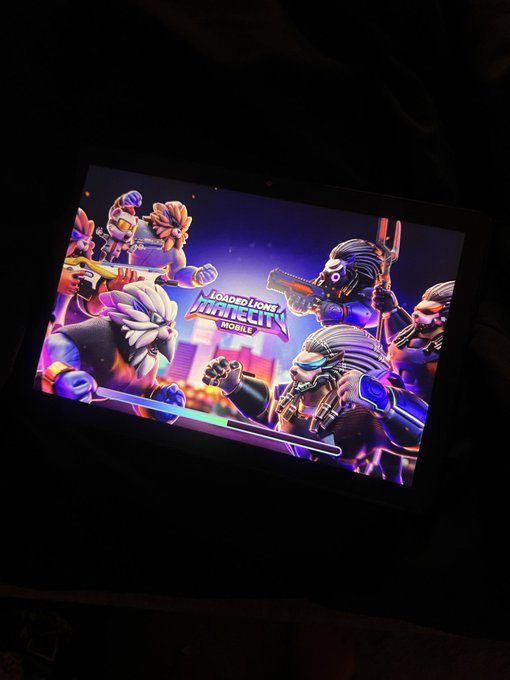 A dimly lit photograph of a mobile device screen displaying the loading screen for 'Loaded Lions: Mane City Mobile,' featuring cartoon lion characters in a vibrant city setting.