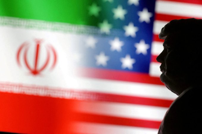A silhouette of a person is shown in profile against a backdrop featuring the Iranian flag and the American flag, visually symbolizing the tensions between the two nations as the U.S. issues escalating threats against Iran's economy and ports.