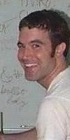 A man in a white shirt smiles while looking back over his shoulder in front of a whiteboard.