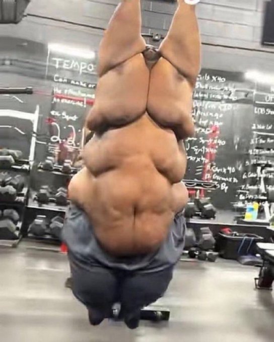 A shirtless bodybuilder displaying extremely large back muscles while doing a lat pulldown exercise in a gym, with a workout schedule board visible in the background.