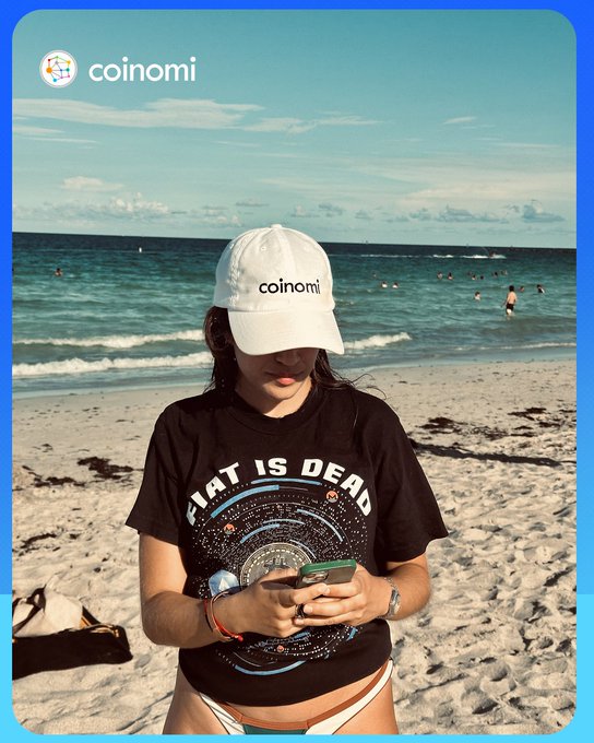 A promotional image showing a woman on a beach wearing a white 'coinomi' cap and a 'FIAT IS DEAD' t-shirt, looking at her phone, with the Coinomi logo in the top left.