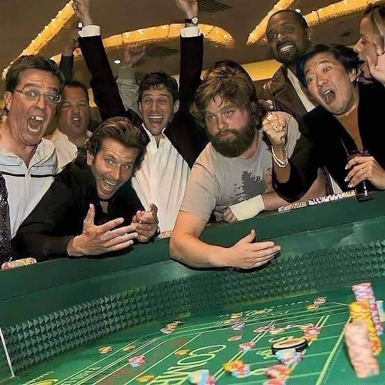 A group of excited, cheering men crowd around a green felt casino table with chips and betting numbers visible, capturing the chaotic thrill of gambling. The image illustrates the "crypto events marketing in 2026" post by humorously comparing the high-stakes, speculative frenzy of crypto promotion to a rowdy casino gamble.