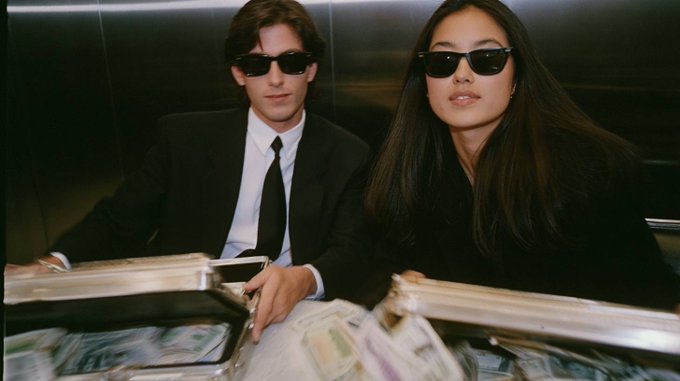 A man and woman in black suits and sunglasses pose with silver briefcases overflowing with stacks of cash. The image's imagery of luxury, wealth, and high-stakes spending complements the post's message about maximizing returns through cashback trading rewards with the $HYPE ticker.