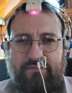 Selfie of a bearded man wearing glasses and a complex head-mounted device with wires and a red light positioned on his forehead.