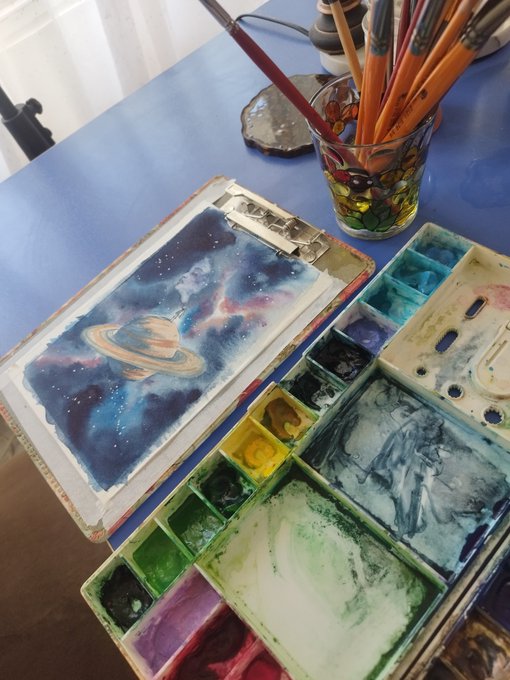 A blue desk holds a watercolor painting of a cosmic scene featuring Saturn, an open palette box with various paint colors, a glass of paintbrushes, and a clipboard.