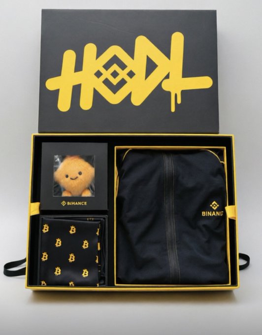 A black and gold Binance branded gift box containing a small plush toy, a black garment with the Binance logo, and a patterned cloth accessory.