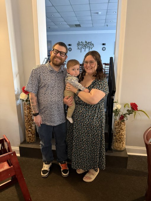 A family portrait of a man, a woman holding a baby, and a toddler standing together in a doorway decorated with floral arrangements.