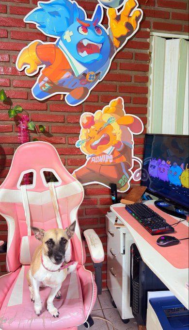 A small tan and white dog sits on a pink and white gaming chair in a cozy gaming setup, with a desktop computer displaying a purple Axie Infinity character visible on the right, and two colorful Axie-style character wall cutouts—one blue labeled "HD" and one orange labeled "ADRIAN"—decorating the exposed brick wall behind, perfectly matching the post's celebration of an Axie Infinity-loving gamer pup.