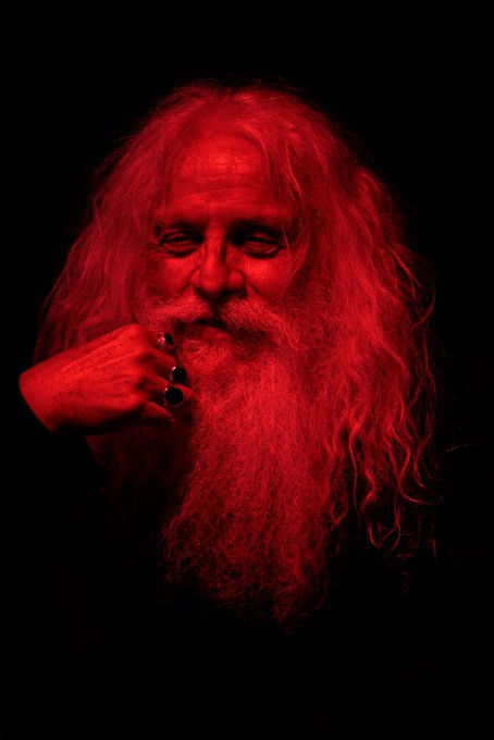 A dramatic portrait of an elderly man with long white hair and beard illuminated in red lighting, wearing rings on his fingers.