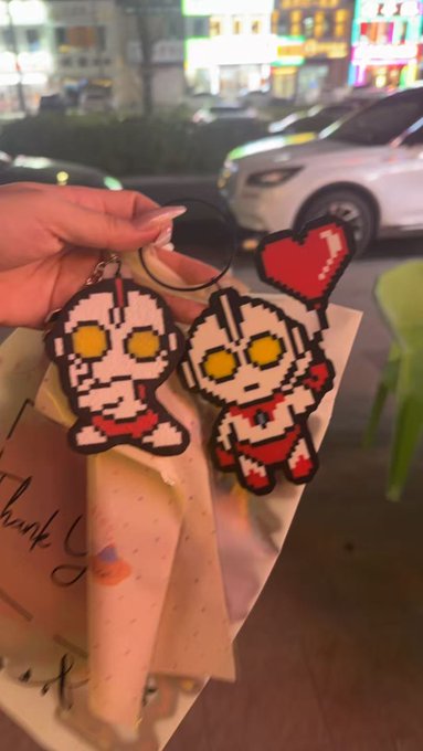 A hand holds two pixel-art Ultraman keychains, one with a red heart, against a nighttime street background with a parked car.