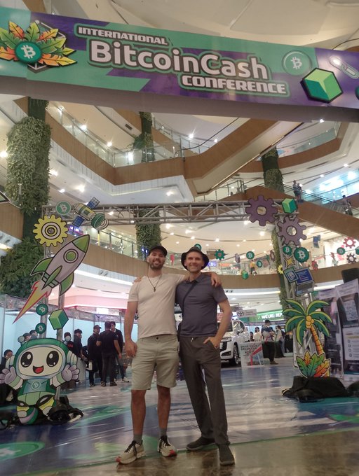 Two men posing together under a banner reading 'INTERNATIONAL BitcoinCash CONFERENCE' at a mall venue decorated with crypto-themed cutouts.