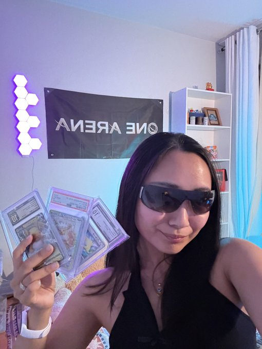 A selfie of a woman wearing sunglasses holding up several graded Pokémon trading cards in plastic sleeves, with an 'ONE ARENA' banner and hexagonal LED lights visible in the background.