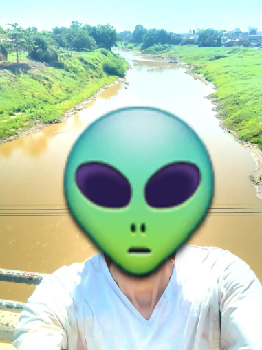 A person in a white shirt sitting in front of a muddy river with green banks, their face completely obscured by a large green alien emoji overlay.