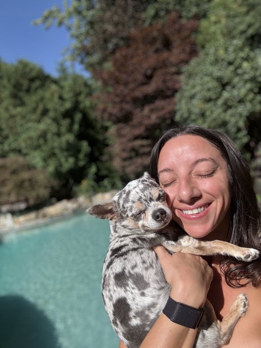 A smiling woman with dark hair holds a sleeping spotted puppy close to her face in front of a pool surrounded by lush trees.