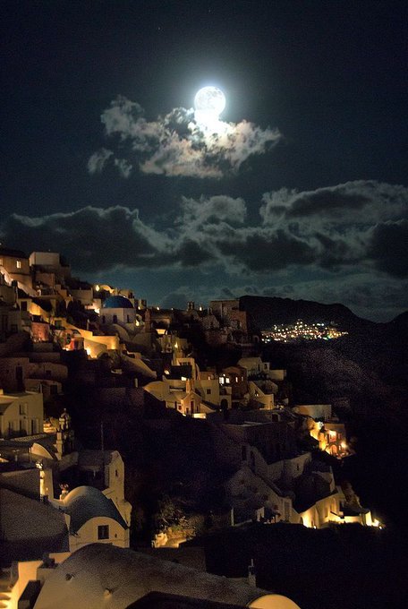 A nighttime photograph shows a hillside village with illuminated buildings under a bright full moon and scattered clouds.
