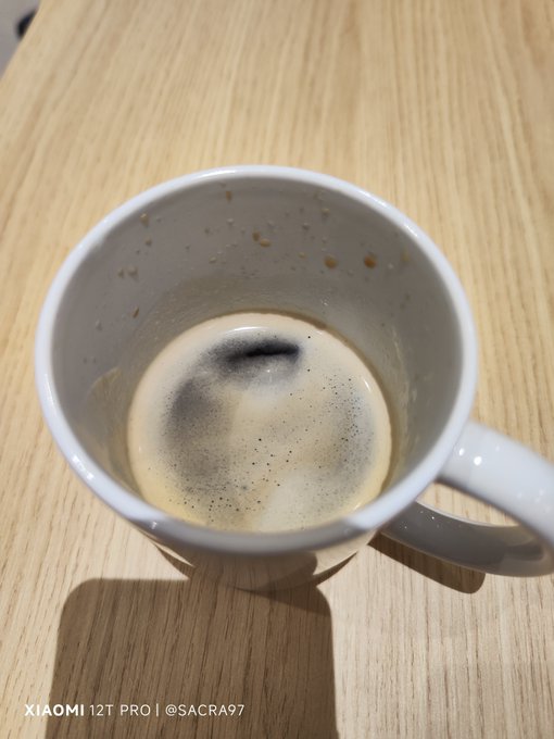 A white ceramic mug sits on a wooden table, containing a frothy beverage with a swirl of dark coffee or chocolate powder on top, with a watermark reading 'XIAOMI 12T PRO | @SACRA97'.
