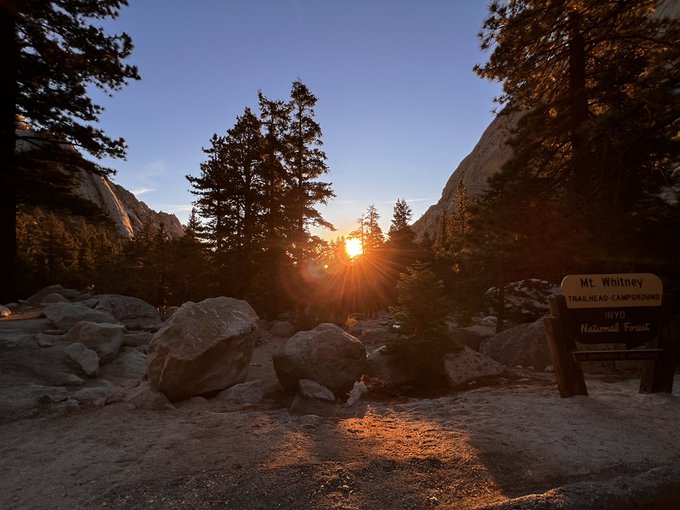 A scenic sunset view at the Mt. Whitney Trailhead in Inyo National Forest, with golden light streaming through pine trees onto a rocky path beside a brown trailhead sign.