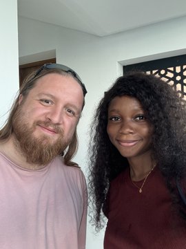 Two people pose closely together for a selfie, with the man on the left having long blonde hair, a beard, sunglasses perched on his head, and a light pink shirt, while the woman on the right has long curly dark hair, a nose ring, a delicate necklace, and a maroon top. The image captures a warm, friendly meeting that aligns with the post about IslandDAO, where the author expresses excitement about connecting with online friends in person—likely including the man pictured—while enjoying a Brazilian BBQ.