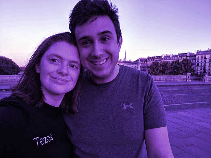 The image features two people, a man and a woman, standing close together with their arms around each other's shoulders. They are both smiling for the camera, creating an endearing moment captured in time. The background of the photo shows buildings, suggesting that they might be in a city or urban setting. The caption to the image reads: "La historia que no te contaron de... Kathleen y Arthur Breitman - un matrimonio joven que dejo Wall Street y Google para crear su propia blockchain". This phrase is likely an inside joke between the couple, and it adds a personal touch to the photo.