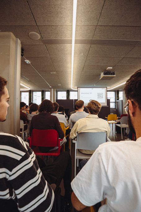 The back view of an audience seated in a modern conference room with acoustic ceiling panels, watching a presentation with a QR code on the projector screen.