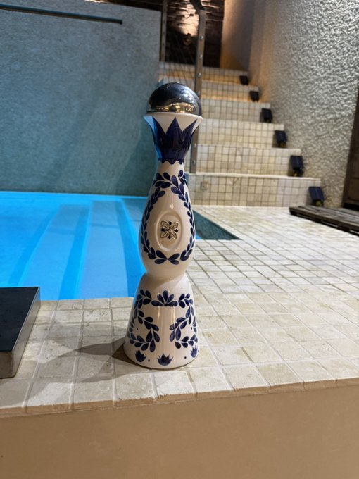 A bottle of Clase Azul tequila, with its signature blue and white floral patterned ceramic decanter and dark blue cap, sits poolside near the water's edge, perfectly matching the caption's "adult hydration break" toast.