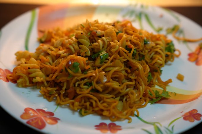 The image shows a plate of noodles, possibly ramen or another type of pasta dish, sitting on top of a table. The noodles are covered with various toppings such as vegetables and sauce, making for an appetizing meal. The plate is placed on a white plate adorned with colorful flowers, adding to the visual appeal of the dish. The image also contains text that reads "G Afternoon fam... Taking a little time out for lunch. This short break is much needed in the middle of work. What'S on your menu this afternoon? #Wallchain #LunchBreak #GoodAfternoon".