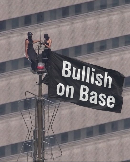 Two people in black attire climb atop a radio/communications tower in a downtown setting, unfurling a large black flag that reads "Bullish on Base" in white text—visually echoing the post's declaration with a literal display of the phrase.