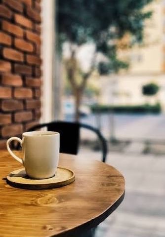 A white ceramic coffee cup on a saucer sitting on a round wooden café table, with a blurred street and brick building in the background.