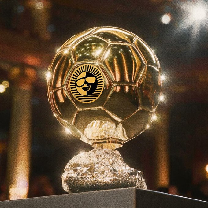 A close-up, warmly lit photograph of a golden Ballon d'Or trophy, featuring a football atop a crystal base with a sun emblem and face design on the ball.