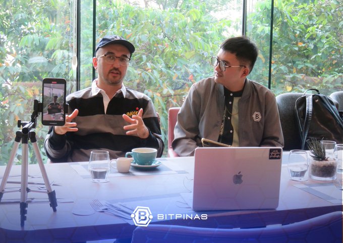 Two men seated at a table during what appears to be a podcast or interview for BitPinas, with a phone on a tripod recording them and an Apple laptop in front.